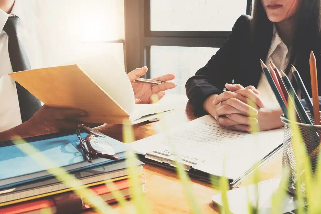 Two attorneys talking with paperwork and notebooks on a desk.