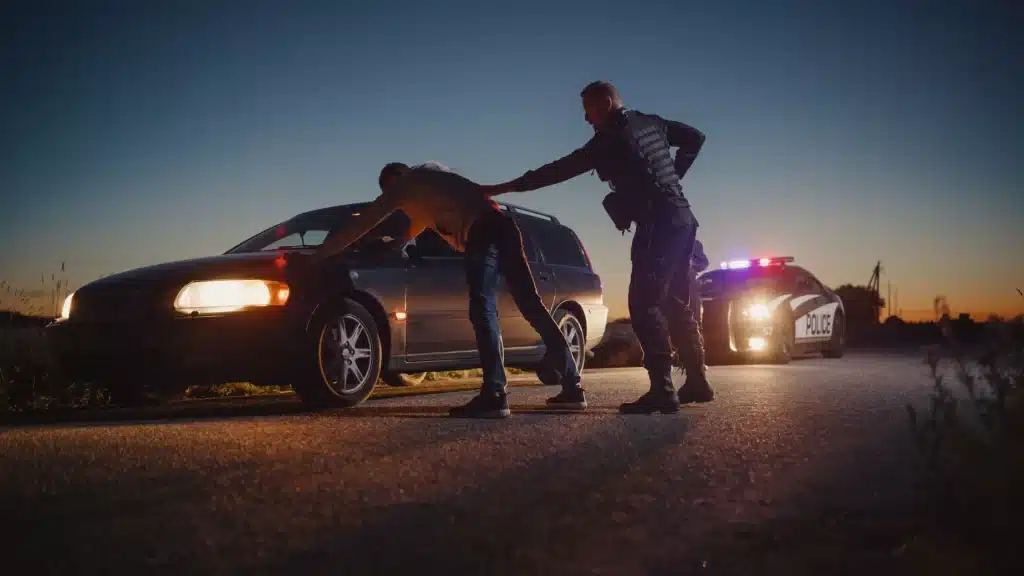 Police officer conducting a search of a driver during a traffic stop at night, with a police car’s flashing lights illuminating the scene.